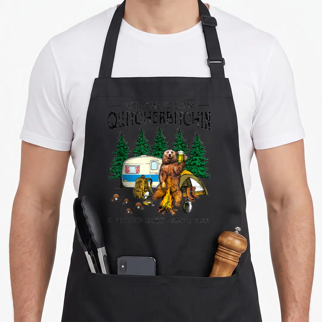 Welcome beer apron - Cartoon bear holding beer mug near campfire with camper trailer and pine trees — shown on a black cotton canvas apron against a white background