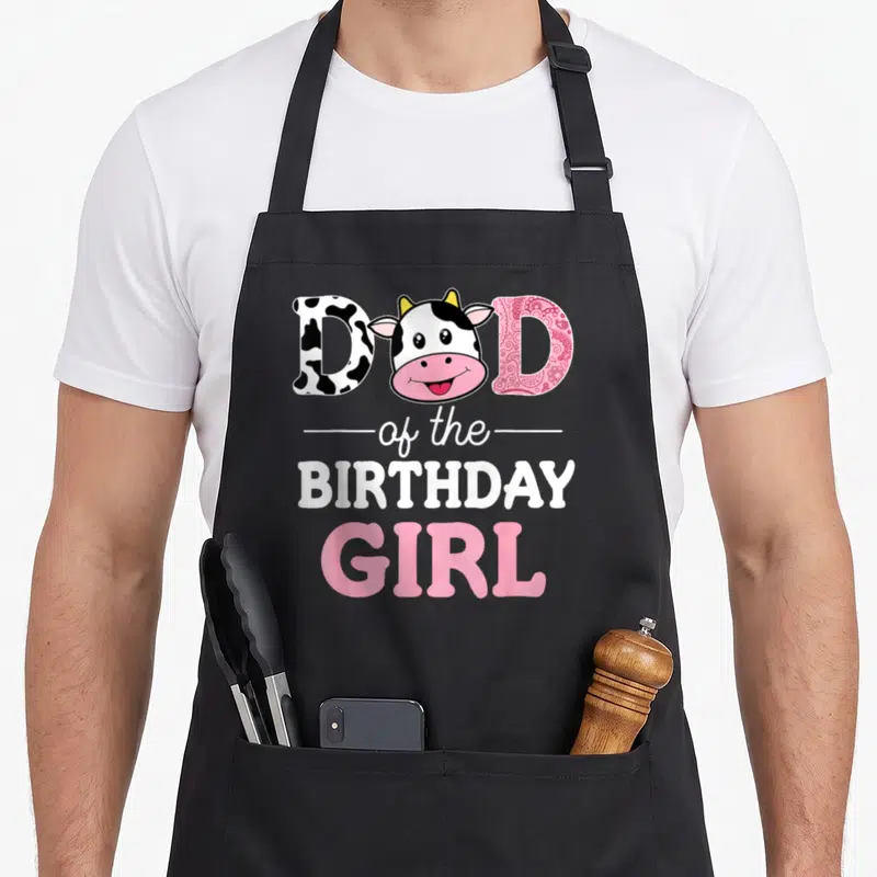 Farm grill daddy apron - Cartoon cow head replacing letter A in patterned DAD of the Birthday Girl text — shown on a black cotton canvas apron against a white background