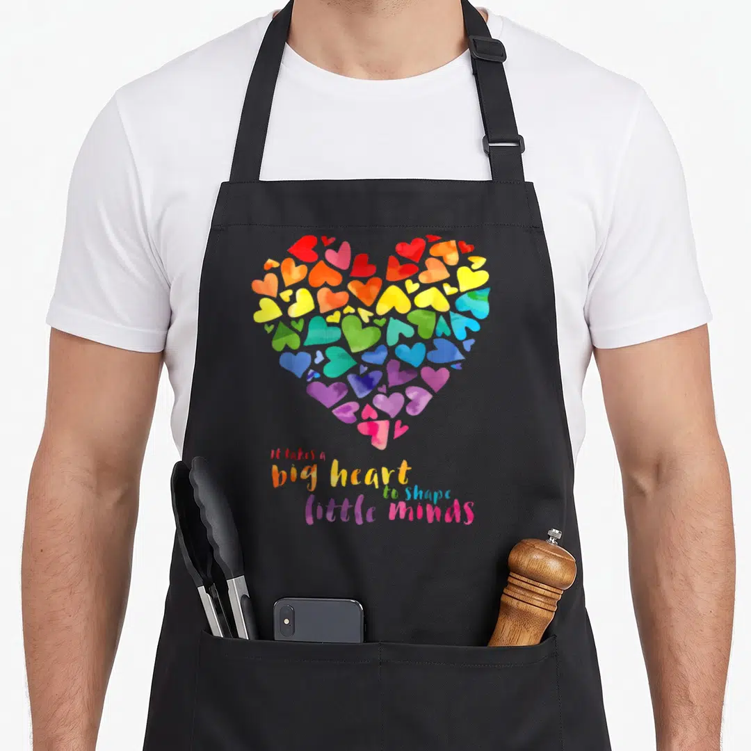 Shape teacher apron - Rainbow watercolor heart composed of smaller hearts above rainbow text reading it takes a big — shown on a black cotton canvas apron against a white background