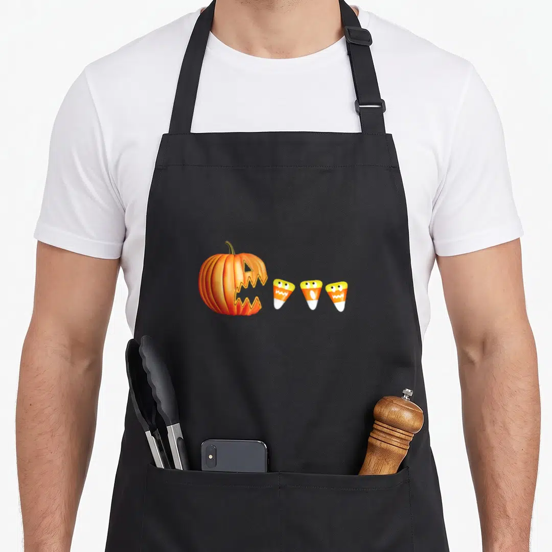 Lantern eating halloween apron - Cartoon pumpkin with open mouth facing three candy corn characters with eyes — shown on a black cotton canvas apron against a white background