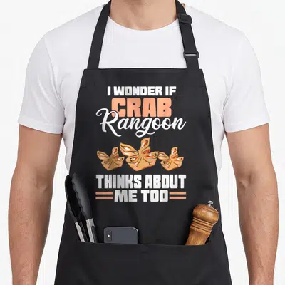 Wonder crab apron - I Wonder If Crab Rangoon Thinks About Me Too text surrounding three dumplings in peach and brown — shown on a black cotton canvas apron against a white background