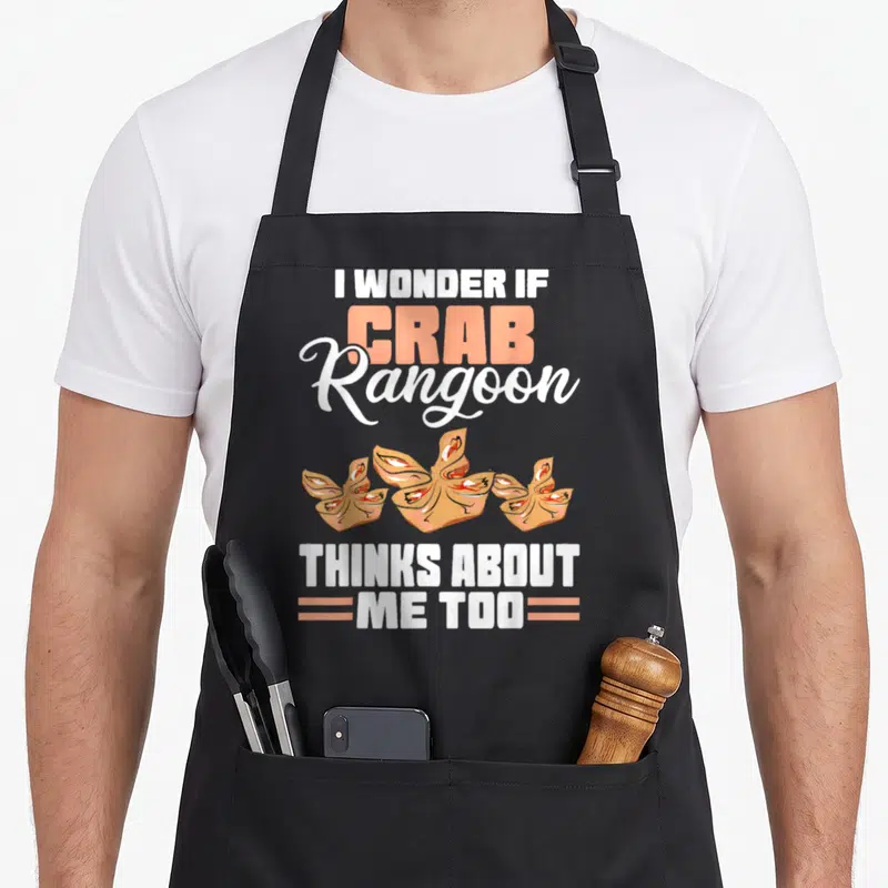 Wonder crab apron - I Wonder If Crab Rangoon Thinks About Me Too text surrounding three dumplings in peach and brown — shown on a black cotton canvas apron against a white background