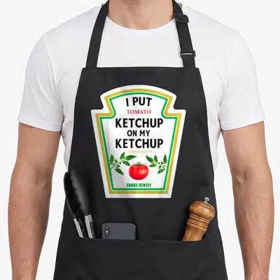 Ketchup mustard apron - Ketchup bottle label print reading I Put Tomato Ketchup on My Ketchup with tomato illustration — shown on a black cotton canvas apron against a white background