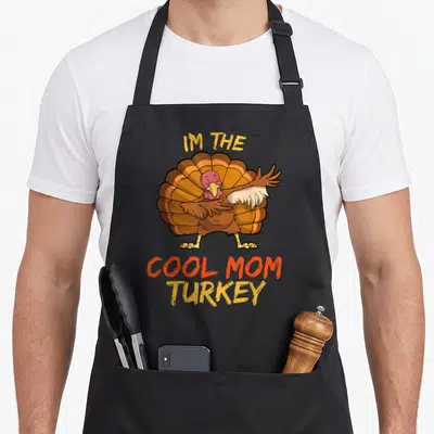 Funny family funny turkey apron - Cartoon turkey with IM THE COOL MOM TURKEY text in brown orange yellow lettering — shown on a black cotton canvas apron against a white background