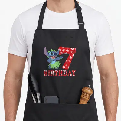 7th stitch apron - Blue hula character next to red floral 7 and BIRTHDAY text — shown on a black cotton canvas apron against a white background