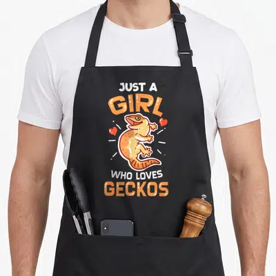 Lizard lounge apron - Cartoon tan gecko between text just a girl who loves geckos with red hearts — shown on a black cotton canvas apron against a white background