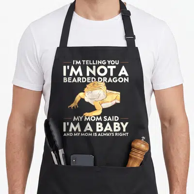Lizard bearded dragon apron - Cartoon orange and yellow bearded dragon framed by white text about mom saying it is — shown on a black cotton canvas apron against a white background