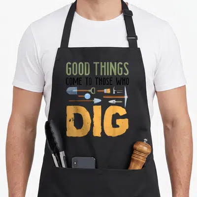 Archaeologist antique apron - Good Things Come To Those Who Dig text with shovels and pickaxes in orange and green — shown on a black cotton canvas apron against a white background