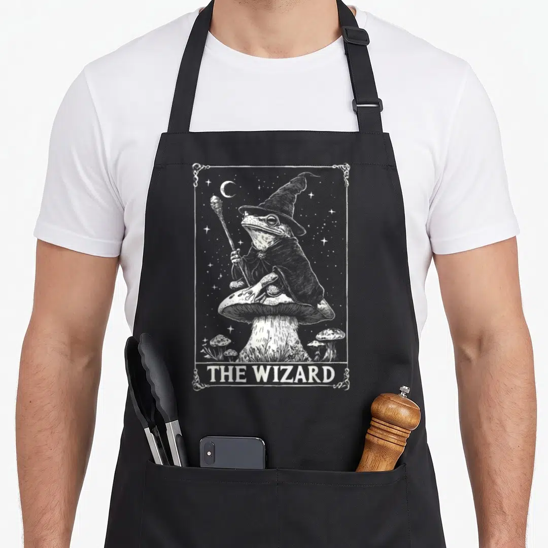 Tarot cottagecore apron - Frog wizard holding staff on mushroom with THE WIZARD text and starry border — shown on a black cotton canvas apron against a white background