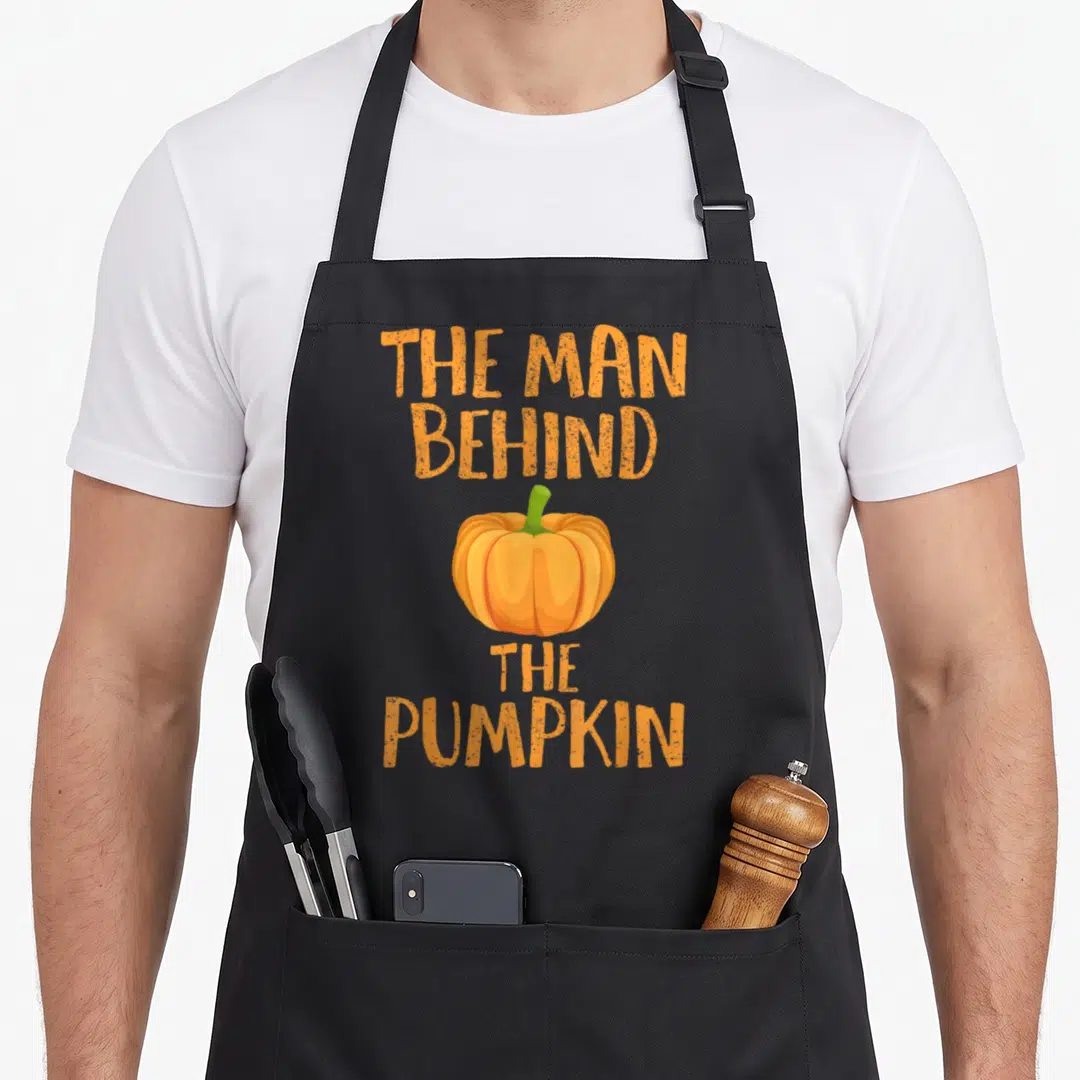 Pregnancy cute halloween aprons - Orange text reading The Man Behind The Pumpkin surrounding a central orange pumpkin — shown on a black cotton canvas apron against a white background