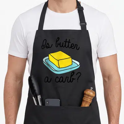 Mean butter apron - Is butter a carb text surrounding a cartoon yellow butter block on a light blue dish — shown on a black cotton canvas apron against a white background
