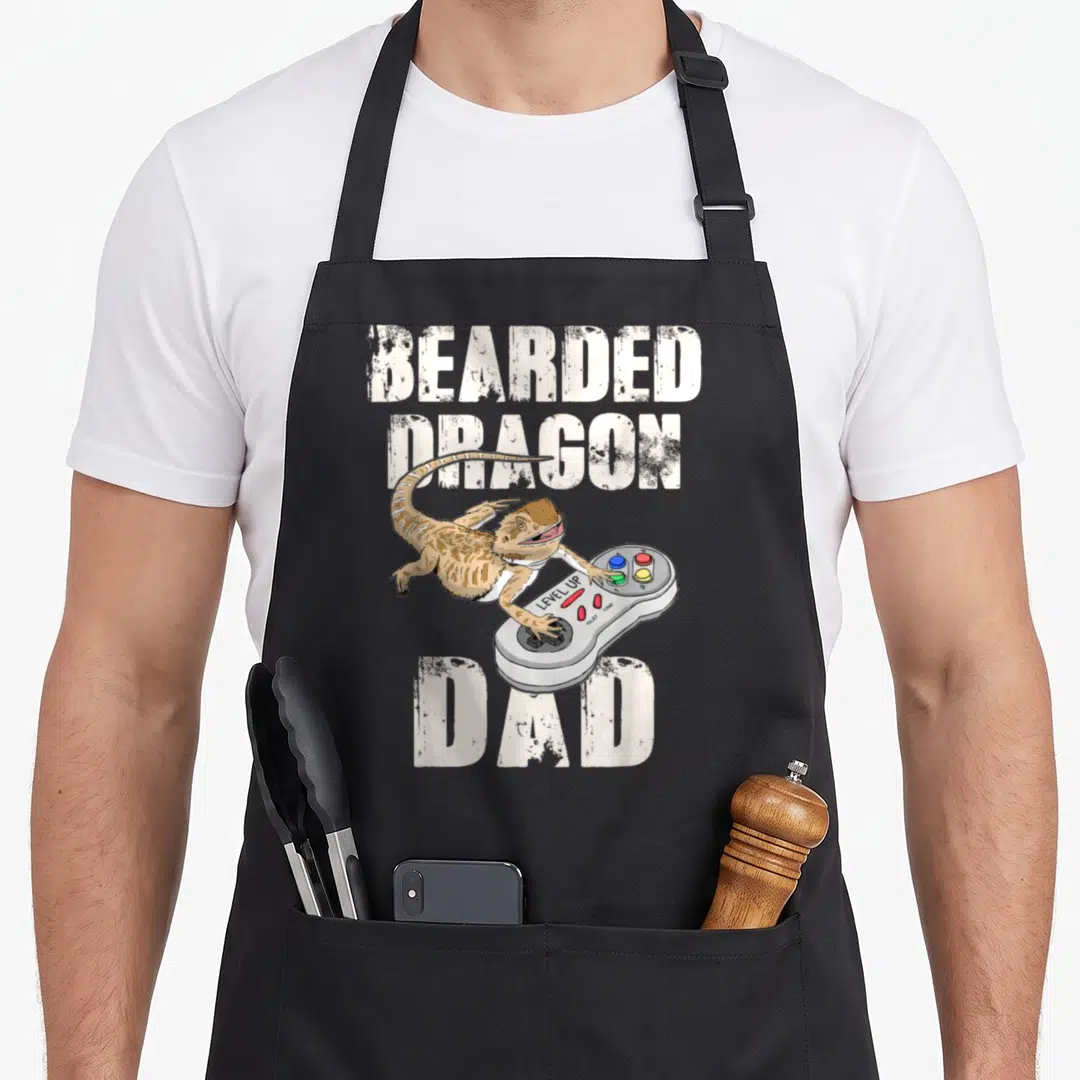 Reptile video game apron - Cartoon bearded dragon holding grey controller with LEVEL UP text between large white words — shown on a black cotton canvas apron against a white background