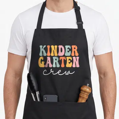 Groovy crew apron - Stacked retro lettering reading KINDER GARTEN crew in pastel colors — shown on a black cotton canvas apron against a white background