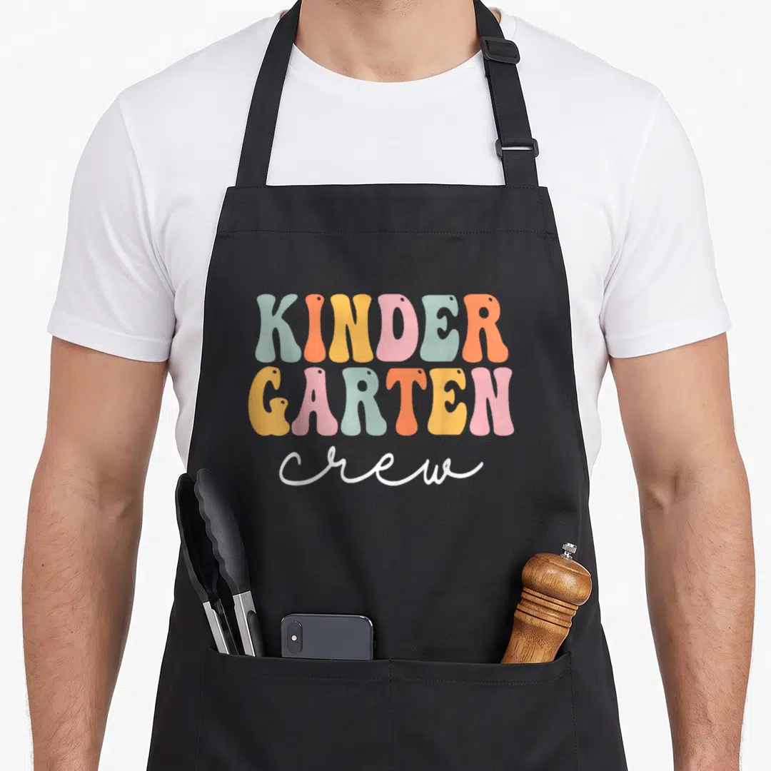 Groovy crew apron - Stacked retro lettering reading KINDER GARTEN crew in pastel colors — shown on a black cotton canvas apron against a white background