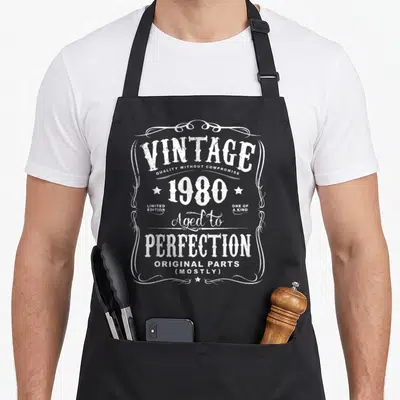 38th birthday apron - White distressed vintage 1980 text stacked inside ornamental frame with stars — shown on a black cotton canvas apron against a white background