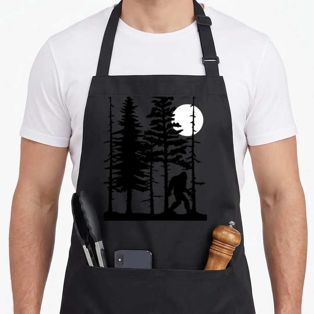 Hiding forest bigfoot apron - Black silhouette of a bigfoot walking past pine trees with a white moon behind them — shown on a black cotton canvas apron against a white background