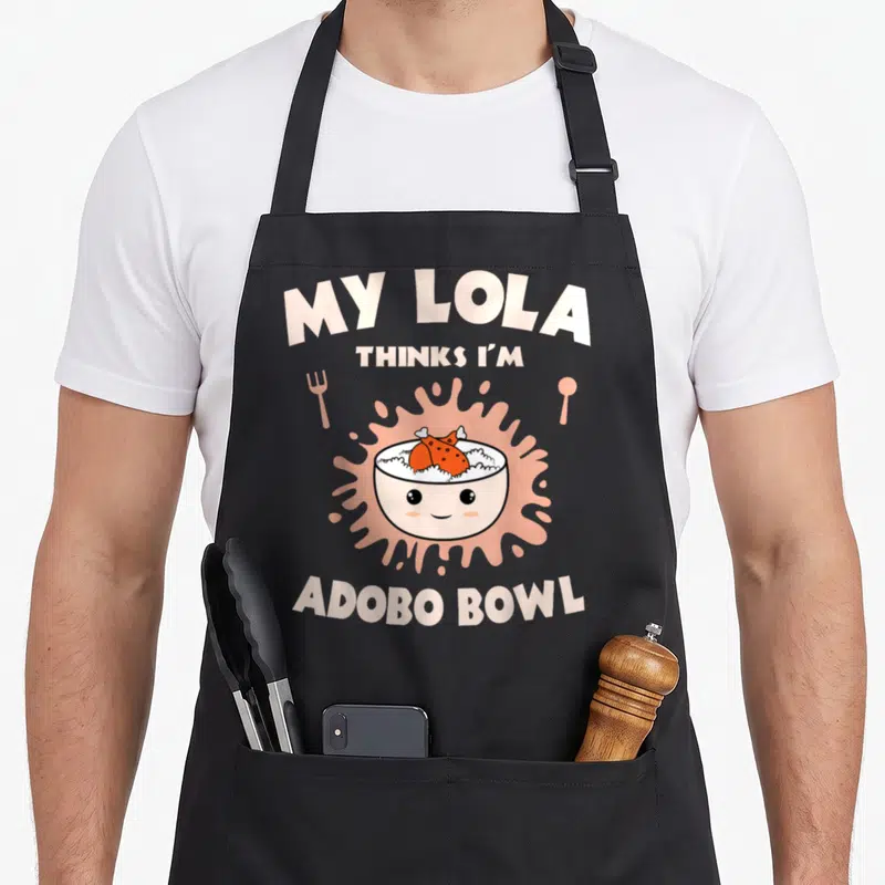 Adobo chicken apron - Cartoon rice bowl with face and adobo chicken on pink splatter with fork and spoon — shown on a black cotton canvas apron against a white background