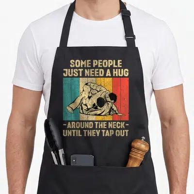 Hug jitsu apron - Two grapplers on beige teal yellow orange red black stripes with sarcastic hug tap out text — shown on a black cotton canvas apron against a white background