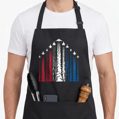 Show 4th of july apron - White jet planes in V-formation leaving vertical red, white, and blue smoke trails on a gray — shown on a black cotton canvas apron against a white background