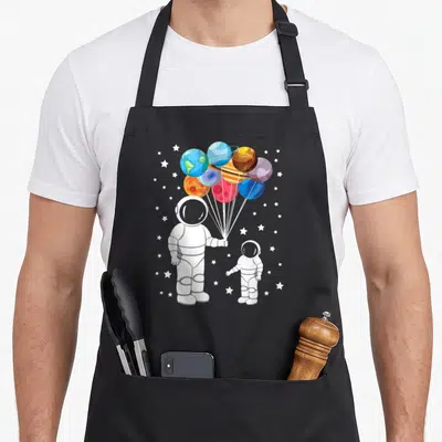 Astronaut solar system apron - Two white cartoon astronauts holding colorful planet balloons surrounded by scattered — shown on a black cotton canvas apron against a white background