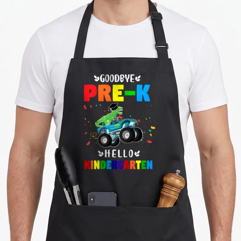 Pre hello apron - Dino in cap on monster truck with goodbye pre-k hello kindergarten text and confetti — shown on a black cotton canvas apron against a white background