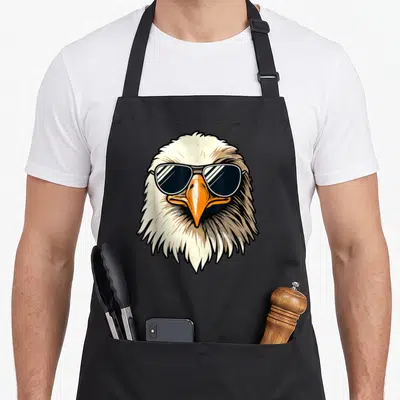 Funny bald 4th of july apron - Cartoon eagle head wearing dark sunglasses with orange beak and white feathers — shown on a black cotton canvas apron against a white background