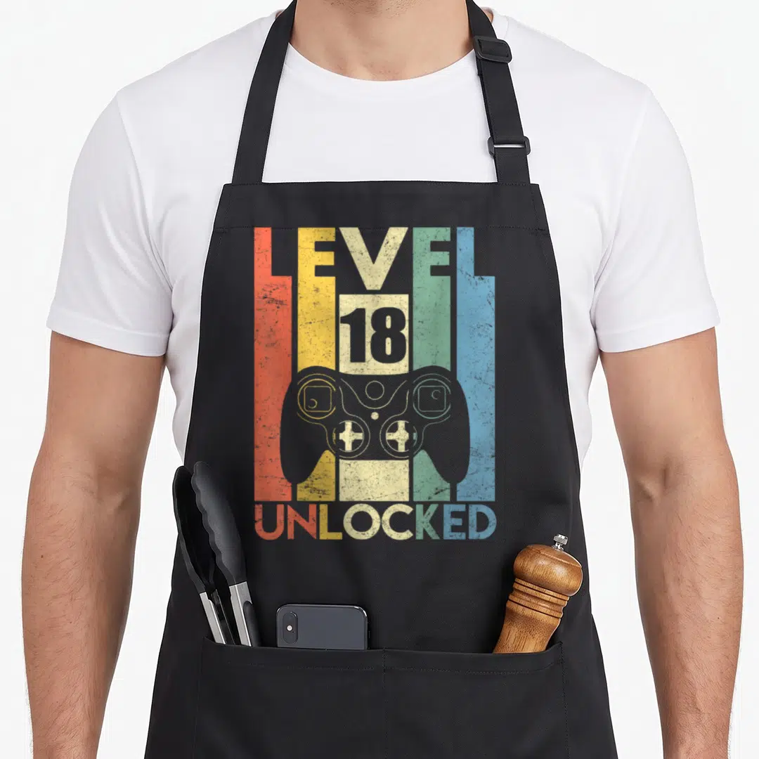 Complet birthday apron - Retro game controller with Level 18 Unlocked text on red yellow cream green blue stripes — shown on a black cotton canvas apron against a white background