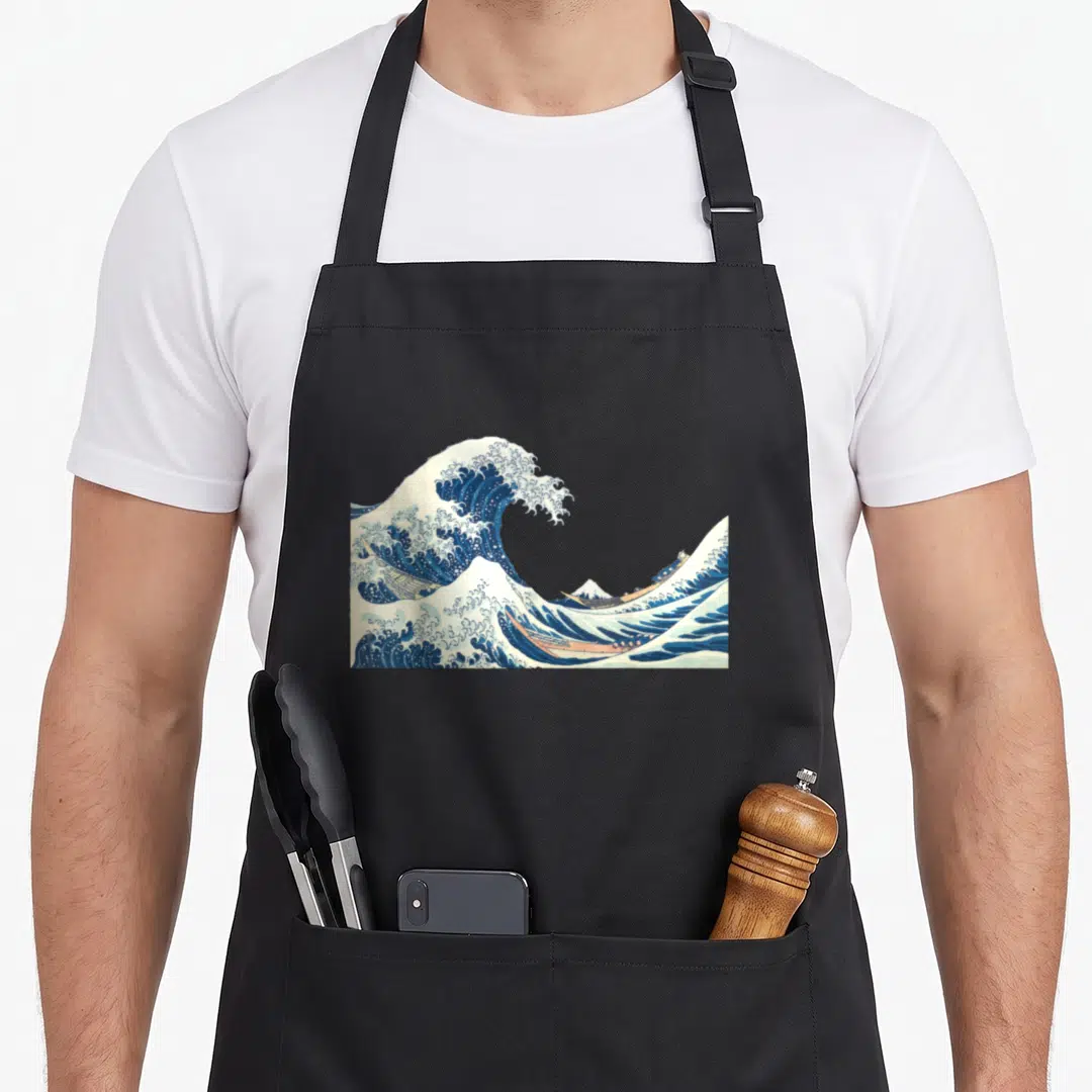Katsushika wave apron - Blue and white wave with three tan boats and a small mountain in realistic Japanese style — shown on a black cotton canvas apron against a white background