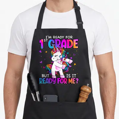 Ready grade unicorn apron - Cartoon unicorn dabbing surrounded by I'M READY FOR 1ST GRADE text and scattered hearts — shown on a black cotton canvas apron against a white background