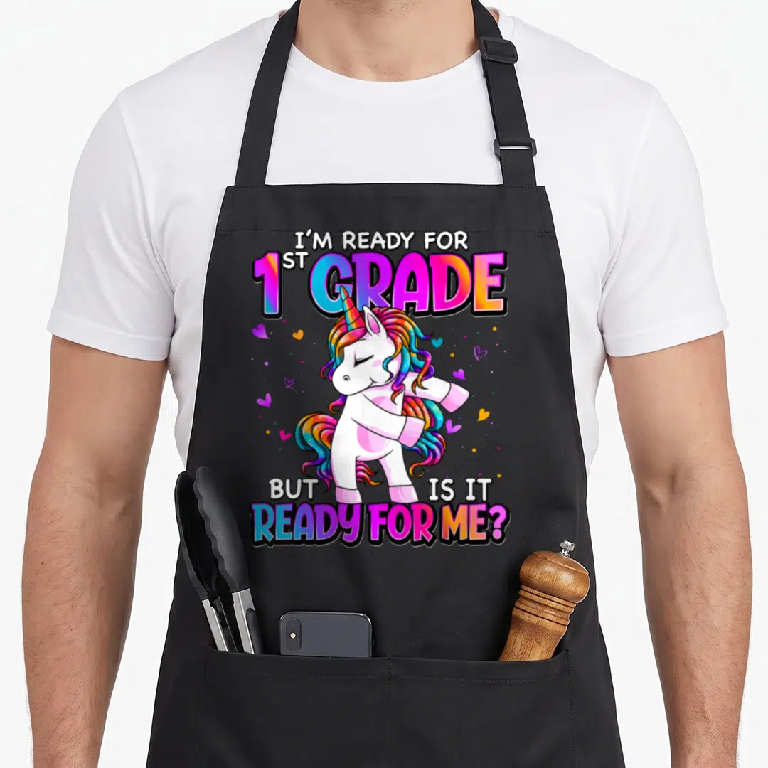 Ready grade unicorn apron - Cartoon unicorn dabbing surrounded by I'M READY FOR 1ST GRADE text and scattered hearts — shown on a black cotton canvas apron against a white background