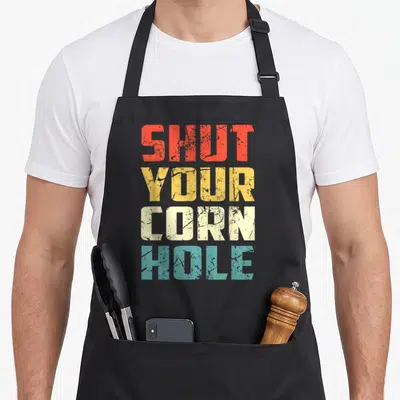 Bean bags and aprons - Stacked retro text Shut Your Cornhole in red yellow cream and teal distressed font — shown on a black cotton canvas apron against a white background