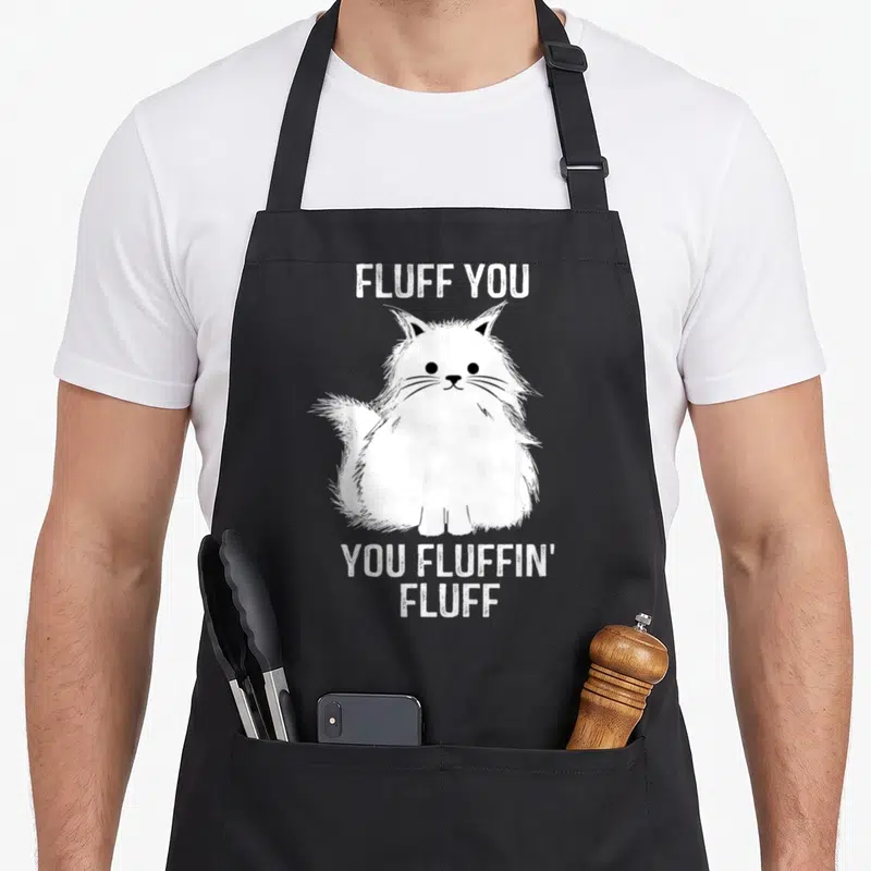 Fluff fluffin kitten apron - White cartoon cat between text reading FLUFF YOU and YOU FLUFFIN' FLUFF on grey background — shown on a black cotton canvas apron against a white background
