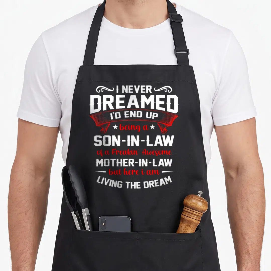 Dreamed funny son in law apron - White and red typography reading I never dreamed I'd end up being a son in law — shown on a black cotton canvas apron against a white background