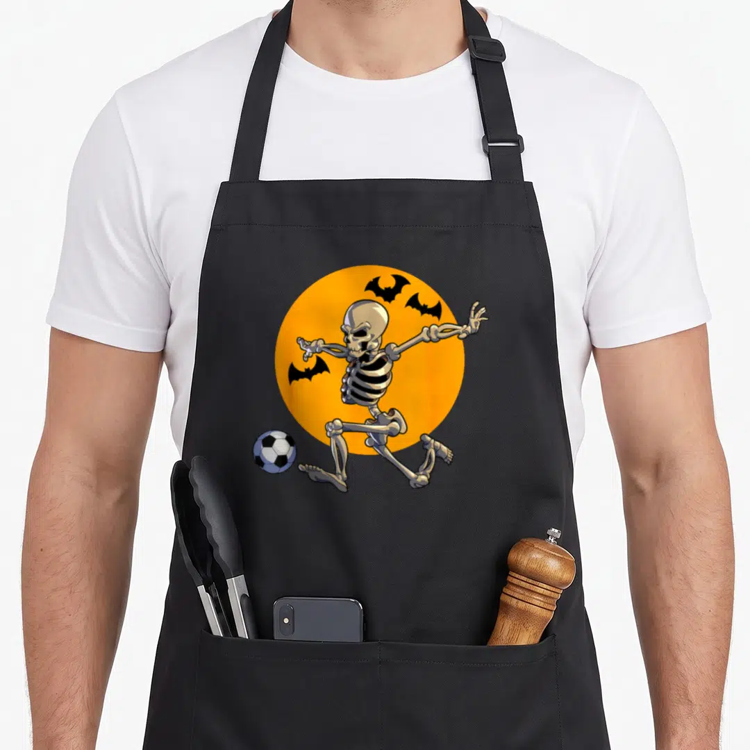 Soccer halloween apron - Cartoon skeleton kicking soccer ball in front of orange circle with three bats — shown on a black cotton canvas apron against a white background
