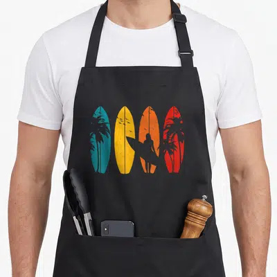 Surf surfing player apron - Four blue yellow orange red surfboards with grey surfer and palm tree silhouettes — shown on a black cotton canvas apron against a white background