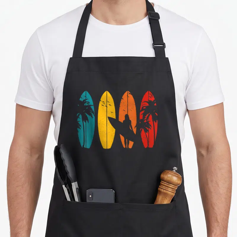Surf surfing player apron - Four blue yellow orange red surfboards with grey surfer and palm tree silhouettes — shown on a black cotton canvas apron against a white background