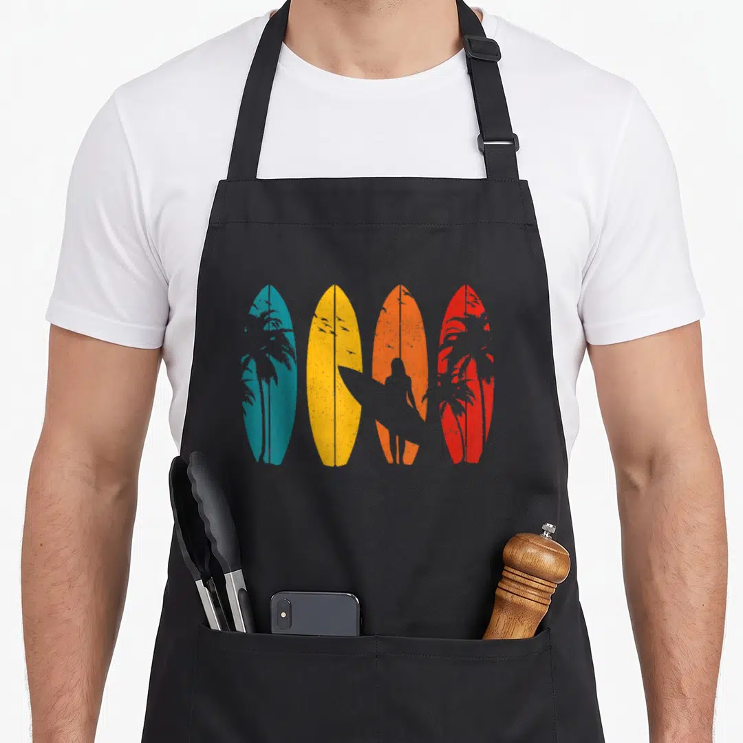 Surf surfing player apron - Four blue yellow orange red surfboards with grey surfer and palm tree silhouettes — shown on a black cotton canvas apron against a white background