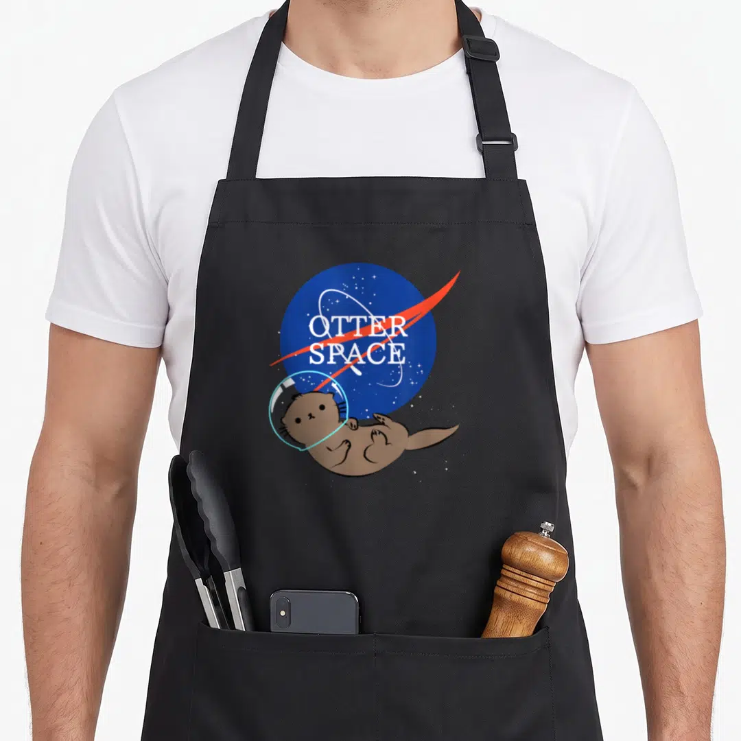 Harry otterhound apron - Brown otter in helmet floating before blue planet with white OTTER SPACE text and red streak — shown on a black cotton canvas apron against a white background