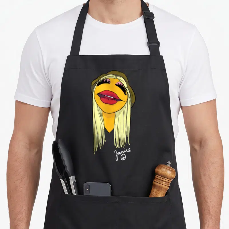 Janice face apron - Cartoon yellow face with blonde hair, olive hat, red lips, name Janice and peace symbol below — shown on a black cotton canvas apron against a white background