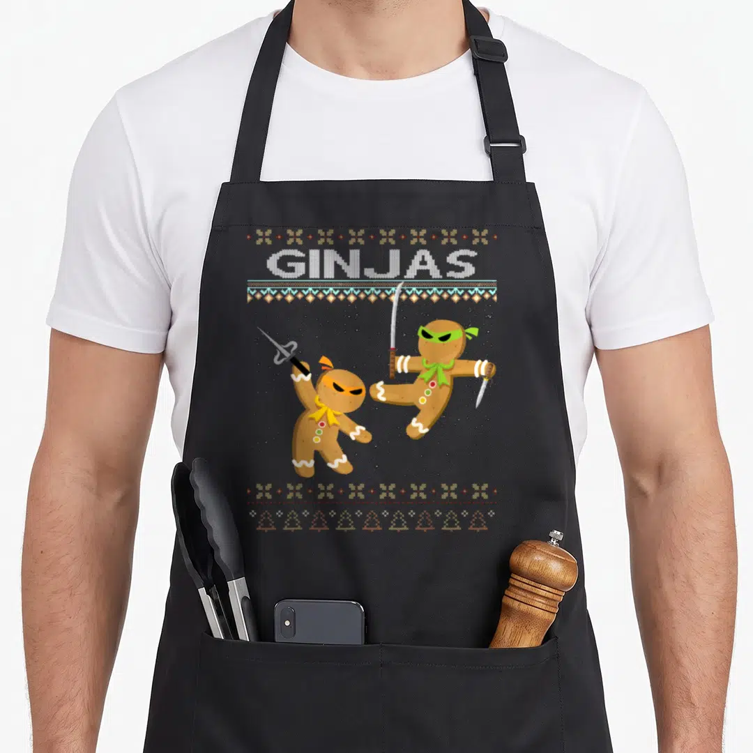Ginjas gingerbread apron - Two ninja gingerbread men fighting under GINJAS text with knitted border patterns — shown on a black cotton canvas apron against a white background