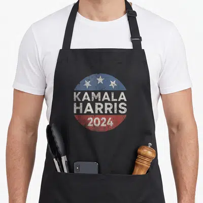Button presidents apron - Distressed circular badge with Kamala Harris 2024 text, three stars, and red white blue bands — shown on a black cotton canvas apron against a white background