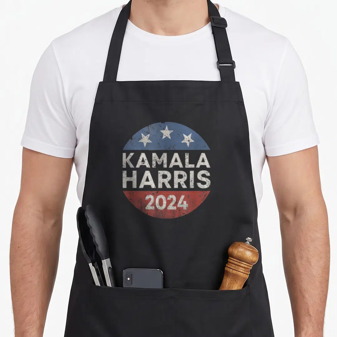 Button presidents apron - Distressed circular badge with Kamala Harris 2024 text, three stars, and red white blue bands — shown on a black cotton canvas apron against a white background