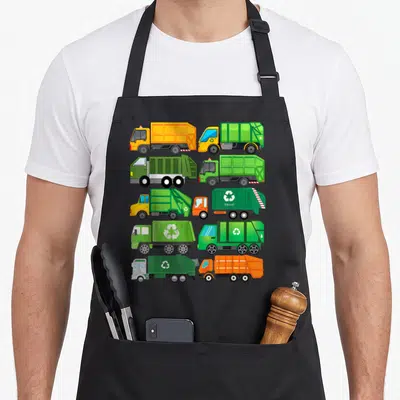 Waste truck apron - Ten cartoon garbage trucks in grid with TRASH text and recycling symbols in green orange yellow — shown on a black cotton canvas apron against a white background
