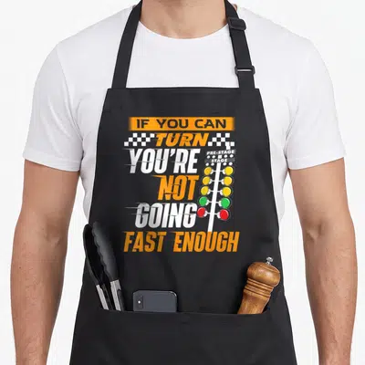 Fast i turn grills on apron - Vertical racing text next to an orange and red Christmas tree light stack with checkered — shown on a black cotton canvas apron against a white background