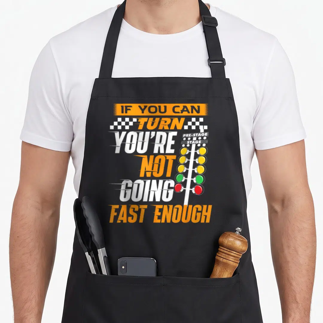 Fast i turn grills on apron - Vertical racing text next to an orange and red Christmas tree light stack with checkered — shown on a black cotton canvas apron against a white background