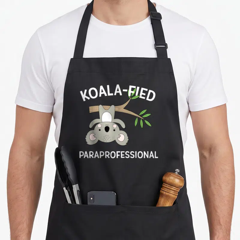 Koala grade flock apron - Cartoon koala hanging from branch with text KOALA-FIED PARAPROFESSIONAL — shown on a black cotton canvas apron against a white background