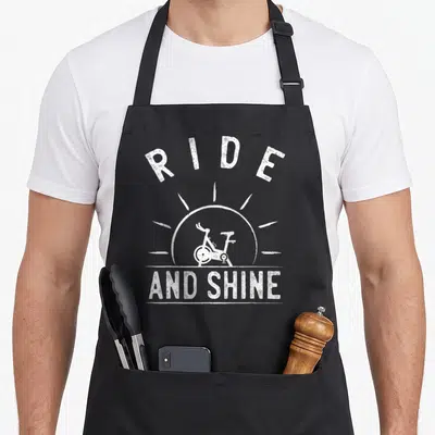 Ride spinning apron - White distressed sunburst with stationary bike and text RIDE AND SHINE — shown on a black cotton canvas apron against a white background