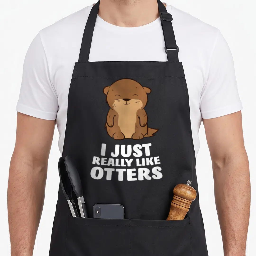 Cute otter apron - Cartoon brown and white otter sitting above white text reading I JUST REALLY LIKE OTTERS — shown on a black cotton canvas apron against a white background