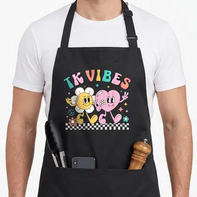 Groovy daisy teacher apron - TK VIBES text over flower and heart characters on checkerboard with stars and peace sign — shown on a black cotton canvas apron against a white background