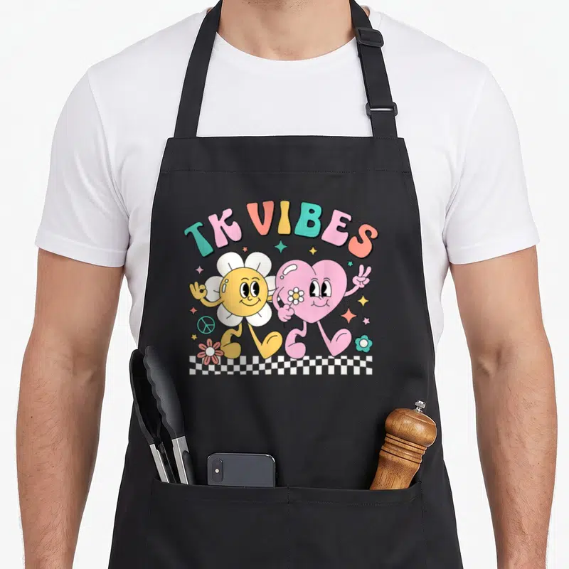 Groovy daisy teacher apron - TK VIBES text over flower and heart characters on checkerboard with stars and peace sign — shown on a black cotton canvas apron against a white background